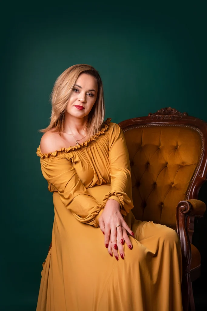 Kobieta w musztardowej sukni siedząca na stylowym fotelu, patrzy spokojnie w stronę widza.
